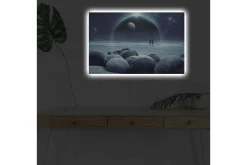 Canvastavla med LED-belysning - Stjärnklädd himmel med två silhuetter vid en strand och stora stenar i förgrunden